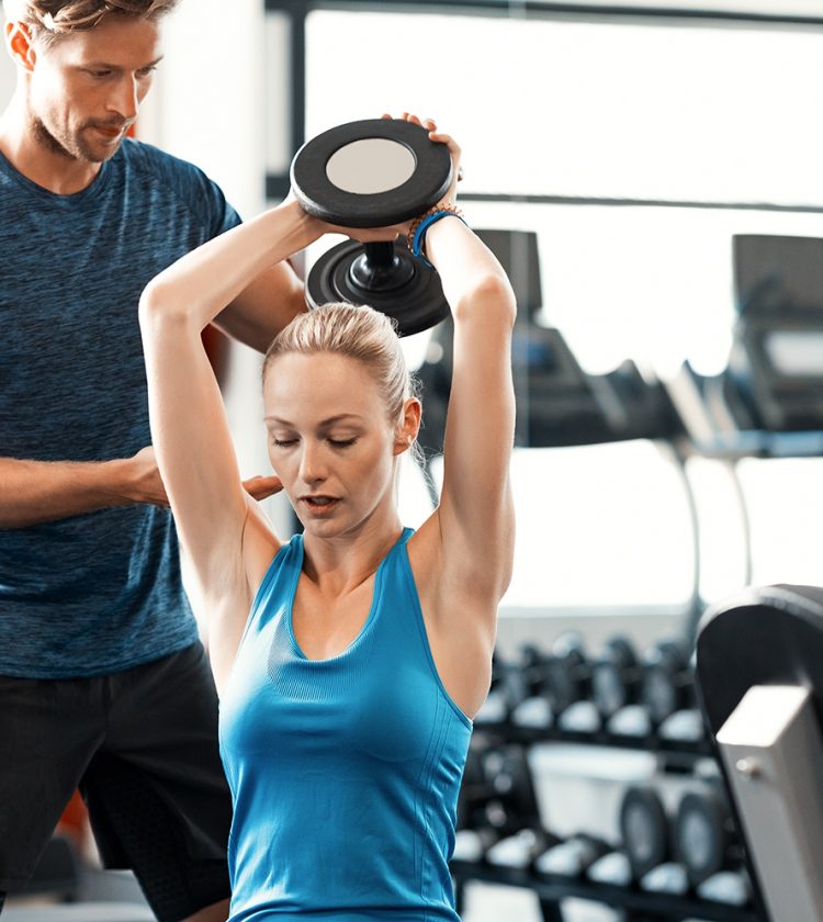 Trainer helping athletic woman at gym. Personal trainer giving weightlifting training to girl in gym. Young woman working out at gym using dumbbells with help of personal trainer.