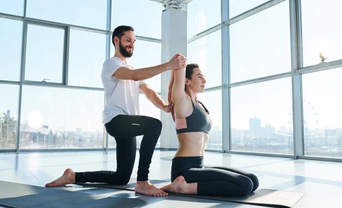 Professional sports trainer helping young active woman to do yoga exercise for back during training in gym